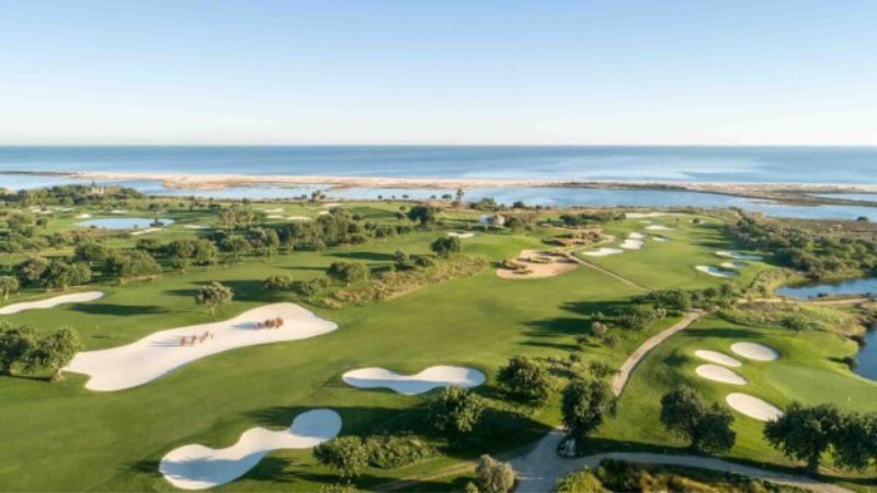 Golfbana i Algarve med fairways, vita bunkrar, vatten och havsutsikt i bakgrunden.